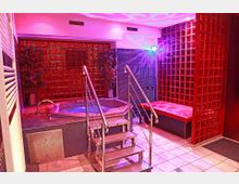 Ein Wellnessraum mit einem Whirlpool, roten Glasbausteinwänden und stimmungsvoller Beleuchtung in pink-lila Tönen. Im Vordergrund befinden sich metallene Treppenstufen, daneben eine gepolsterte rote Sitzbank.