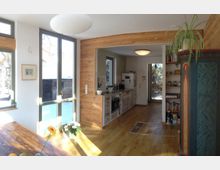 Helle Küche und Essbereich mit großen Fenstern, die viel Tageslicht hereinlassen. Holzelemente an Wänden und Boden sowie offene Regale und schlichte Küchenmöbel prägen den Raum.