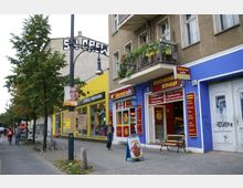 Straßenszene mit kleinen Geschäften in einem Wohngebiet. Ein Gebäude in Gelb und Blau beherbergt einen Getränkeshop und ein Internetcafé, während ein Balkon mit Blumen geschmückt ist; daneben stehen Bäume und ein Gehweg mit Fahrrädern und Fußgängern.