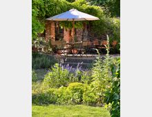 Ein gemütlicher Außenbereich mit Holztischen und -stühlen unter einem großen Sonnenschirm, umgeben von üppigem Grün und blühenden Pflanzen. Im Hintergrund befindet sich eine rustikale Mauer aus roten Ziegelsteinen und Natursteinen, teilweise bewachsen mit Kletterpflanzen.