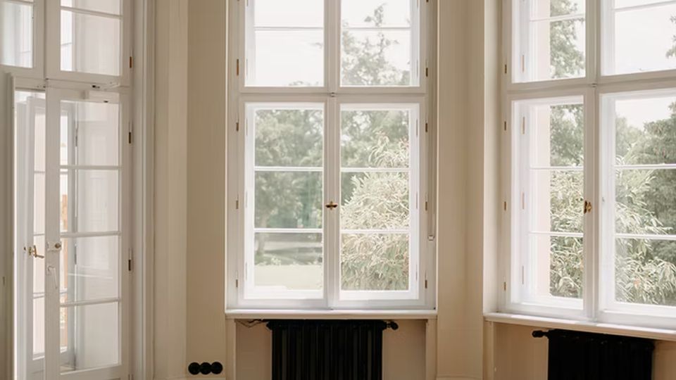 Heller, leerer Raum mit hohen, weißen Sprossenfenstern in einer Erkerfront, durch die Tageslicht auf einen glänzenden Parkettboden mit Muster fällt. An der Decke hängt eine dekorative Pendelleuchte; unter den Fenstern sind schwarze Heizkörper und links eine verglaste Tür.