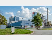 Industrieanlage mit blauen und grauen Gebäuden, hohen Schornsteinen und metallischen Konstruktionen, umgeben von einem Zaun und einer Straße im Vordergrund. Links ein Baum und Grünflächen, rechts vereinzelte Laternenmasten.