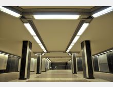 Unterirdische Bahnhofshalle mit großflächigem, beigem Deckenbereich, rechteckigen Leuchtstofflampen, dunklen Säulen mit metallischen Elementen und einem Boden aus hellen und dunklen Fliesen. Die Wände sind mit dunklen Paneelen verkleidet, und im Hintergrund sind Informationsschilder und Rolltreppen erkennbar.