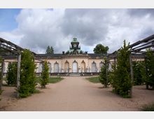 Barockgebäude mit heller Fassade, verziert mit Skulpturen und einem zentralen Uhrturm mit grünlicher Kuppel. Im Vordergrund führen Sandwege durch eine symmetrisch gestaltete Gartenanlage mit Laubbögen.