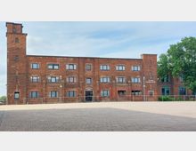 Backsteinbau im rechteckigen, mehrstöckigen Stil mit symmetrischen Fenstern, einem Haupteingang mit der Aufschrift "Hotel" und einem seitlichen Turm. Rechts befindet sich ein Bereich mit der Beschriftung "Uferrum Strandbar & Café", umgeben von gepflastertem Vorplatz und einigen Bäumen.