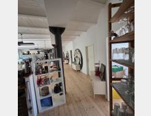 Ein heller Innenraum mit Holzboden und gewölbter Decke, entlang dessen eine offene Küche, Regale mit Haushaltsgegenständen und eine Ausstellungswand mit Bildern und Notizen zu sehen sind. Im Vordergrund stehen zwei Regale, rechts mit Gläsern und links mit Dekoartikeln.