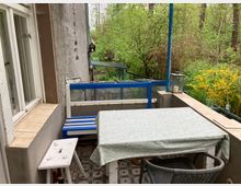 Ein Balkon mit Fliesenboden und leicht verwitterten Möbeln, darunter ein Tisch mit gemusterter Tischdecke, ein Holzstuhl und eine Bank mit blauen Elementen. Im Hintergrund dichter grüner Bewuchs und Büsche.