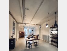 Loftähnlicher Raum mit industriellem Charakter, weiß verputzten Wänden und freiliegenden Deckenbalken. Im Vordergrund stehen ein Holztisch mit Hockern und im Hintergrund sind Sitzmöbel, Regale, Lampen und ein großer Fensterbereich sichtbar.