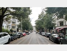 Straßenszene mit beidseitig parkenden Autos und Straßenbahnschienen in der Mitte, flankiert von mehrstöckigen Wohngebäuden mit Balkonen und Geschäften im Erdgeschoss; Bäume säumen den Weg und schaffen eine grüne Allee. Im Hintergrund ist ein weißes Gebäude sichtbar.