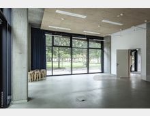 Moderner, minimalistischer Innenraum mit Betonwänden, hellem Boden und einer großen Fensterfront, die den Blick auf einen parkähnlichen Außenbereich mit Bäumen freigibt. Im Raum stehen mehrere gestapelte Holzstühle, und eine Tür führt in einen weiteren Bereich.