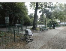 Kleiner Park mit Sandwegen, grünen Bänken und schmiedeeisernen Gittern. Im Vordergrund steht ein Kinderwagen, im Hintergrund ist eine Person an einer Bank zu sehen, umgeben von Bäumen und Büschen.