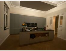 Moderne Küche mit grauen Einbauschränken, einer zentralen Kücheninsel mit schwarzer Arbeitsplatte und Blick auf eine Tür und ein Fenster. Die Decke ist mit Stuck dekoriert, und der Raum ist schwach beleuchtet.