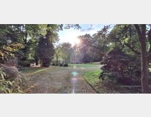 Weitläufiger Park mit breitem, unbefestigtem Weg durch Rasenflächen, umgeben von hohen Laubbäumen, Sträuchern und einer Weide im Hintergrund. Links stehen ein hölzerner Schuppen und große Tonkrüge, während die tief stehende Sonne durch das Blätterdach scheint.