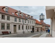 Straßenszene in einer Altstadt mit Fachwerk- und Putzgebäuden, die rote Ziegeldächer haben. Im Hintergrund steht ein Wasserturm mit rundem Fundament und auffälliger Kuppel, während einige Menschen auf dem Gehweg spazieren.