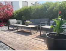 Holzterrasse im Garten mit modernen grauen Loungemöbeln und einem niedrigen Couchtisch. Im Hintergrund dichte grüne Hecke und ein roter Zierbaum, vorne ein großer Pflanzkübel mit hoher Pflanze und orangefarbener Blüte.