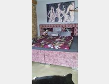 Ein Schlafzimmer mit einem großen, rosafarben gepolsterten Bett, das mit einer dunkel gemusterten Bettdecke dekoriert ist. An der Wand hängt ein abstraktes Gemälde von Figuren, und links ist ein Teil einer freigelegten Backsteinwand sichtbar.