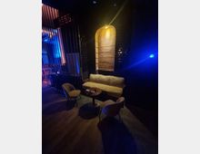 Dunkles, stilvolles Lounge-Interieur mit einem gepolsterten Sofa, zwei Sesseln und einem runden Beistelltisch auf Holzboden. Im Hintergrund ein beleuchteter, gewölbter Wandbereich aus Holz und dekorative Gitterelemente.