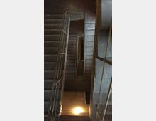 Treppenhaus mit mehreren Etagen in einem Gebäude aus Ziegelstein, mit Metalgeländern und Betonstufen. Der Raum ist schwach beleuchtet, mit einem Bodenlicht auf der untersten Ebene.