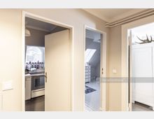 Fluransicht mit drei offenen Türen: links führt eine Tür in eine Küche mit einem gewölbten Fenster und sichtbaren Arbeitsflächen, in der Mitte geht eine Tür in ein Badezimmer mit weißen Fliesen und Oberlichtern, rechts ist ein Raum mit einem weißen Schrank erkennbar.