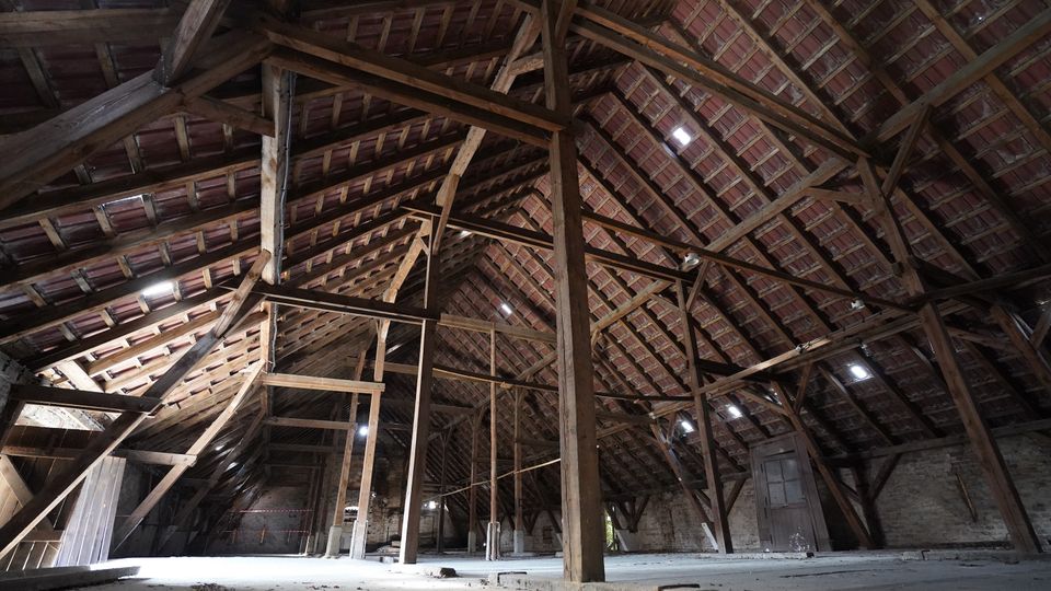 Großer Dachbodenraum mit sichtbarem Holzbalkenwerk und Ziegel-Unterdach, getragen von hohen Stützen und Querstreben. Der Boden ist weitgehend leer, mit gemauerten Außenwänden und mehreren kleinen Dachöffnungen, durch die Licht einfällt.