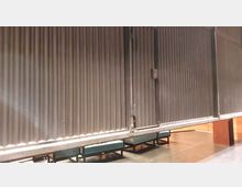 Metallische Wand- oder Trennkonstruktion mit vertikalen Paneelen und sichtbaren Verschlüssen, darunter eine Sitzgelegenheit mit grünen Polstern auf schwarzen Metallrahmen. Der Hintergrund zeigt teilweise Holzverkleidungen und einen dunklen Fußboden.