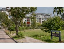 Mehrstöckiges Wohngebäude mit Balkonen und einer teils verglasten Dachfassade im Hintergrund, umgeben von einer Grünanlage mit Rasen, Bäumen und Sträuchern. Rechts im Vordergrund ein Straßenschild mit der Aufschrift „Freiberger Straße 14–45“.