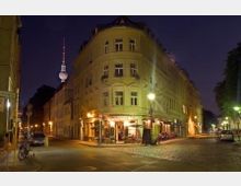 Nachtszene einer Straßenecke mit einem beleuchteten historischen Wohngebäude, auf dessen Erdgeschoss sich ein Restaurant befindet. Im Hintergrund ist der beleuchtete Berliner Fernsehturm zu sehen, umgeben von Bäumen und Straßenlaternen.