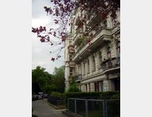 Mehrstöckiges historisches Wohngebäude mit verzierten Balkonen und großen Fenstern, umgeben von Bäumen und einem Gehweg. Im Vordergrund sind ein Zaun und rote Blätter eines Baumes zu sehen, während der Hintergrund eine ruhige Straße mit parkenden Autos zeigt.