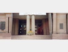 Fassade eines historischen Gebäudes mit breiter Treppe, zwei großen Säulen vor dem Eingang und mehreren Türen. Links und rechts der Säulen sind Gedenktafeln an der sandsteinfarbenen Wand angebracht.