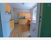 Blick in eine helle Wohnküche mit gelben Einbauküchenschränken, weißem Kühlschrank und Holzboden. Rechts steht ein Tisch mit orangefarbener Tischdecke neben einem großen Fenster; die Wände sind mit gemusterter Tapete und Fliesenspiegel versehen.