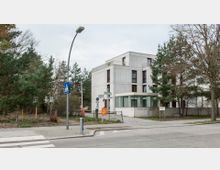 Modernes, rechteckiges Gebäude mit heller Fassade und großen Fenstern, umgeben von Bäumen und Grünflächen. Im Vordergrund eine Straßenkreuzung mit Zebrastreifen, Straßenlaterne, Verkehrsschildern und einem Mülleimer.
