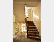Treppenhaus in einem älteren, teils renovierungsbedürftigen Gebäude mit brauner Teppichläufer-Treppe, dunklem Holzgeländer und beigefarbenen Wänden. Große Fenster bringen Tageslicht herein; unten sind ein Heizkörper und eine Zwischenpodestfläche mit Staub und Bauschutt zu sehen.