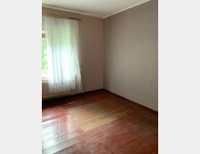 Leerer Raum mit einem braunen Holzboden, einer hellgrauen Wand und einem Fenster mit weißem, halbtransparentem Vorhang. Das Fenster lässt Tageslicht herein, und unter dem Fenster befindet sich ein Heizkörper.