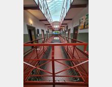 Innenraum eines historischen Gebäudes mit Gefängnisarchitektur, bestehend aus zwei Ebenen mit Zellentüren entlang der Wände und roten Geländern. Oben ist ein Glasdach zu sehen, das Tageslicht in den schmalen Gang leitet.