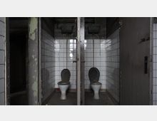 Zwei verlassene Toilettenkabinen mit weißen Fliesenwänden, alten Keramik-Toilettenschüsseln und darüber hängenden Spülkästen. Die Umgebung zeigt Abnutzungs- und Verfallsspuren, wie abblätternde Farbe und Schmutz.