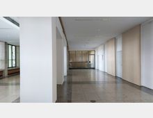 Weißer Flur mit hohen Decken in einem modernen Gebäude, Boden aus polierten Steinfliesen und Wänden aus glatten Paneelen; am Ende des Flurs befindet sich eine Tür aus geriffeltem Glas, flankiert von Fenstern mit Rahmen. Links befinden sich große Fenster mit Blick nach draußen.