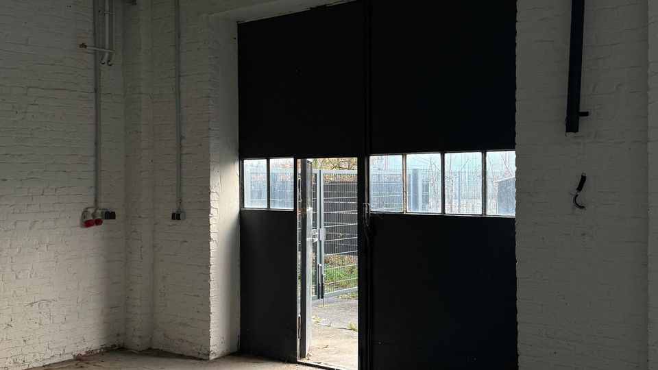 Innenraum einer Industriehalle mit weiß gestrichenen Backsteinwänden und Betonboden; an der Decke verlaufen sichtbare Rohre und eine lange Leuchtstofflampe. Eine große schwarze Schiebetoröffnung mit Fenstersegmenten lässt Tageslicht herein und gibt den Blick nach draußen auf einen eingezäunten Bereich frei.