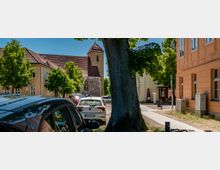 Straßenszene in einem historischen Ortskern mit geparkten Autos, großen Bäumen und niedrigen, hell verputzten Gebäuden mit Ziegeldächern. Im Hintergrund steht eine kleine Kirche mit steinernem Sockel und einem quadratischen Turm mit spitzem Dach.