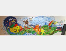 Wandgemälde mit bunten, fantasievollen Elementen, darunter eine riesige gelbe Katze, eine Windmühle, Häuser, eine Eisenbahn, ein Regenbogen und Naturdarstellungen wie Flüsse und Bäume. Der Stil ist verspielt, mit Märchenmotiven und kindgerechten Figuren auf einem weißen Hintergrund.