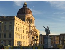 Historisches Gebäude mit einer großen Kuppel und detailreicher Fassade, beleuchtet vom Sonnenlicht, rechts eine Statue auf einem Podest mit einer geflügelten Figur. Im Hintergrund moderneres Gebäude und blauer Himmel.