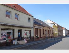 Straßenansicht mit kleinen Wohn- und Geschäftsgebäuden, darunter ein Laden mit großer Schaufensterfront und Eingangsstufen. Die Gebäude haben helle Fassaden, Ziegeldächer und teils geschlossene Rollläden, während eine gepflasterte Straße im Vordergrund verläuft.