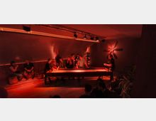 Ein dimm beleuchteter Innenraum mit einer Gruppe von Menschen, die an einem langen Shuffleboard-Tisch spielen. Im Hintergrund sind Pflanzen, warme Wandbeleuchtung und ein großes Wanddekor in Form eines Propellers zu sehen.