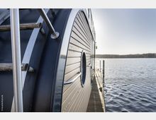 Seitenansicht eines modernen Hausbootes mit Holzverkleidung und rundem Fenster, das an einem Steg auf einem ruhigen Gewässer liegt. Im Hintergrund ist ein bewaldetes Ufer unter klarem Himmel sichtbar.