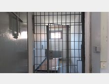 Ein einfacher Gefängnisraum mit einer offenen Metallgittertür, einem kleinen Fenster, das Licht hereinlässt, einem Heizkörper und einem schmalen Bettgestell. Die Wände und der Boden sind schlicht und grau gehalten.