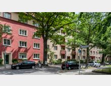 Mehrstöckige Wohngebäude in Rot- und Beigetönen mit weißen Fensterrahmen, umgeben von Bäumen und geparkten Autos entlang einer ruhigen Straße. Ein Straßenschild zeigt die "Ganghoferstraße", und mehrere Fahrräder stehen an einem Fahrradständer.