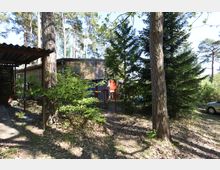 Ein bewaldeter Außenbereich mit einem kleinen Holzgebäude im Hintergrund, umgeben von dichter Vegetation mit Bäumen und Sträuchern. Links im Bild ist ein überdachter Unterstand zu sehen, rechts ein geparkter Pkw.