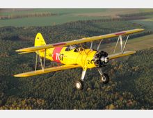 Gelbes Doppeldecker-Propellerflugzeug mit rotem Streifen und der Aufschrift "U.S. Navy" fliegt über eine waldreiche Landschaft, die mit Feldern durchzogen ist. Im Hintergrund sind Bäume und landwirtschaftliche Flächen unter einem klaren Himmel zu sehen.