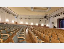 Ein moderner Theatersaal mit Reihen gepolsterter Stühle mit Holzlehnen, einer Bühne auf der rechten Seite und einer hohen, abgerundeten Decke mit integrierter Beleuchtung. An den Wänden befinden sich mehrere Türzugänge und schlichte Wandpaneele mit eingebauten Leuchten.