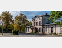 Ein altes, zweigeschossiges Bahnhofsgebäude mit Backsteinverzierungen und teilweise verputzten Fassaden, umgeben von großen Bäumen und einem mit Kopfsteinpflaster ausgelegten Vorplatz. Im Hintergrund ist ein Zaun zu sehen, eingebettet in eine natürliche Umgebung.