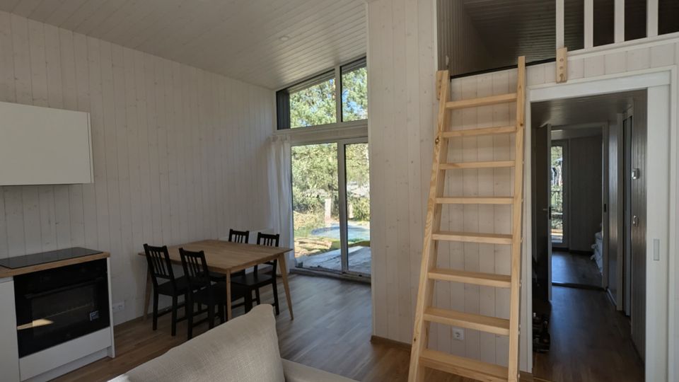 Heller Wohnraum in einem modernen Holzhaus mit hoher Decke, Sofa, kleinem Essbereich und Küchenzeile. Eine Holzleiter führt zu einer offenen Schlaf- oder Galerieebene, daneben große Glas-Schiebetüren mit Blick ins Grüne.